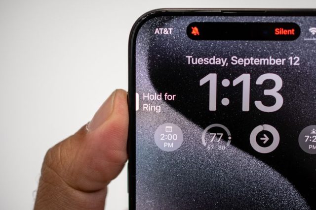 Dari iPhoneIslam.com, Seseorang memegang ponsel dengan jam tangan sambil memperlihatkan tombol aksi baru di iPhone 15 Pro.