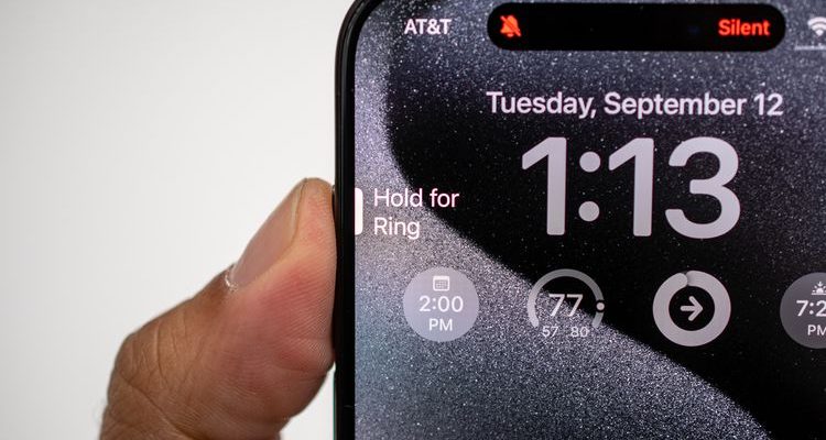 iPhoneIslam.com より、時計を付けた携帯電話を持っている人が、iPhone 15 Pro の新しいアクション ボタンを示しています。