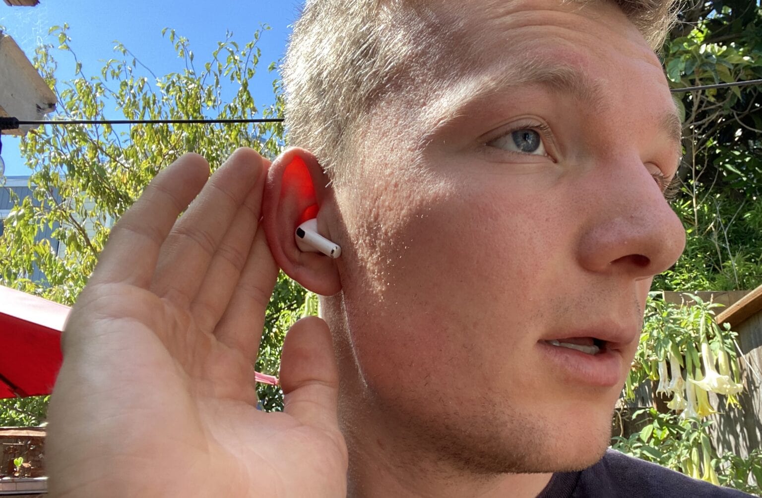 iPhoneIslam.com より、緑の葉が特徴的な日当たりの良い屋外の背景で、若い男性が赤い AirPods 4 を耳に挿入します。