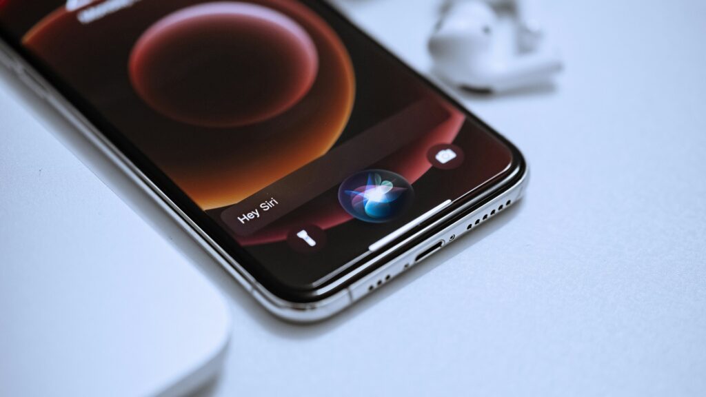 iPhoneIslam.com より、画面上で Siri がアクティベートされ、ライト グレーの表面のワイヤレス イヤフォンの隣にある iPhone スマートフォン。