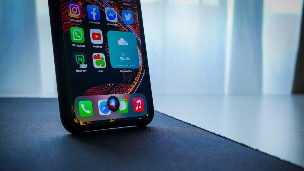 iPhoneIslam.com より、テーブル上のスマートフォンの画面には、薄暗い背景に Facebook、WhatsApp、YouTube などのさまざまなアプリのアイコンが表示されます。