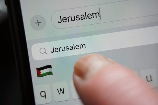 Từ iPhoneIslam.com, hình ảnh cận cảnh một ngón tay đang nhấn nút “Tìm kiếm” trên màn hình điện thoại thông minh có chữ “Jerusalem” được viết và biểu tượng cảm xúc lá cờ Palestine.