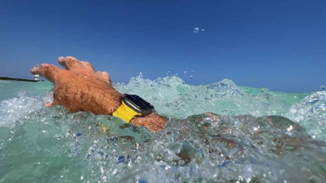 Von iPhoneIslam.com: Nahaufnahme der Hand einer Person, die eine gelbe Apple Watch hält und Wasser in klares, blaues Meerwasser spritzt.