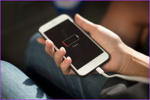 Dari iPhoneIslam.com, Seseorang memegang iPhone dengan indikator baterai lemah di layar, terhubung ke charger, di dalam mobil.