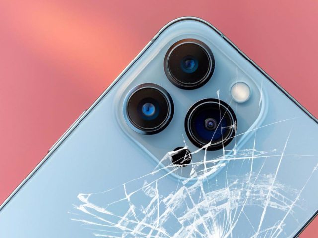 Do iPhoneIslam.com, close-up da câmera traseira de um smartphone com três lentes, uma delas rachada, com processadores M4, em um fundo rosa e azul.