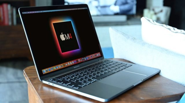 Do iPhoneIslam.com, um laptop em uma mesa de madeira exibindo um gráfico colorido com as letras "cma" na tela, alimentado por processadores M4, em uma sala com um sofá e um fundo desfocado