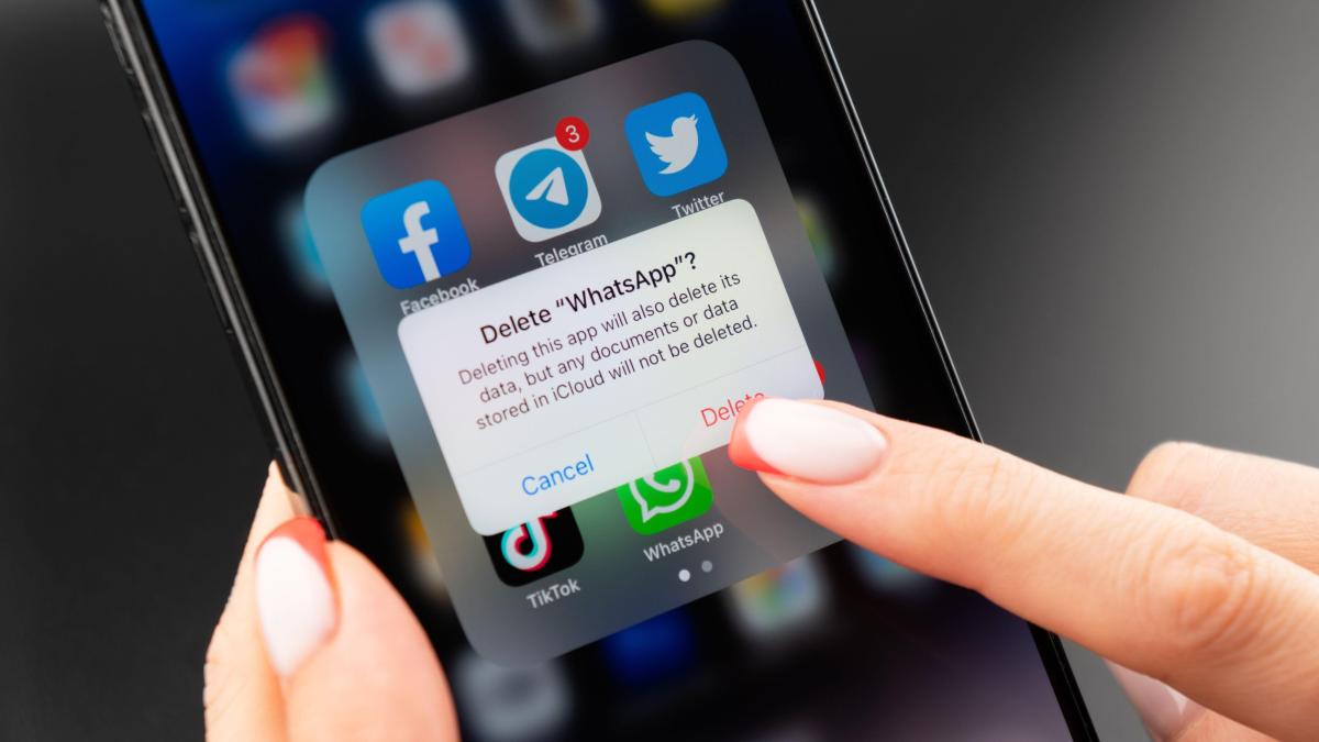 Do iPhoneIslam.com, um close de uma mão segurando um smartphone mostrando um pop-up de confirmação de exclusão do WhatsApp em abril, com outros ícones de mídia social em segundo plano.
