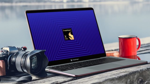 Von iPhoneIslam.com, ein Laptop mit dem Qualcomm Snapdragon-Logo auf dem Bildschirm, platziert auf einem Holzsockel neben einer roten Tasse und einer DSLR-Kamera, mit einem See im Hintergrund.
