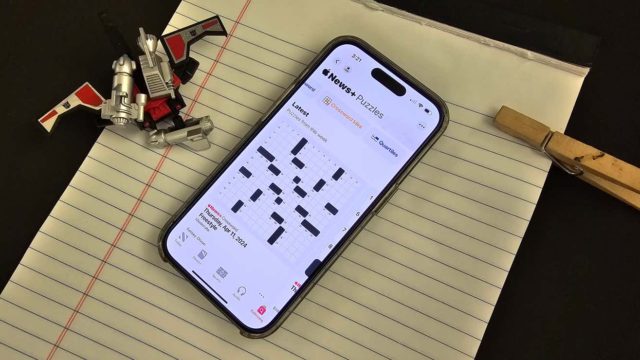 iPhoneIslam.com에서 가져온 달력 앱을 표시하는 스마트폰은 검은색 표면에 장난감 로봇과 나무 빨래집게 옆 줄이 쳐진 노트북 위에 놓여 있습니다.