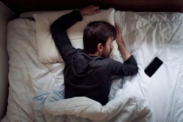 来自 iPhoneIslam.com，一名男子躺在床上，用手臂遮住眼睛，旁边放着一部 iPhone。床上用品是白色的，他似乎正在休息或沮丧。