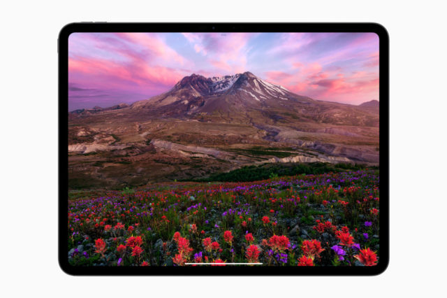 من iPhoneIslam.com، جهاز لوحي رقمي يعرض منظرًا طبيعيًا نابضًا بالحياة مع جبل وزهور برية ملونة تحت سماء غروب الشمس المثيرة في حدث إطلاق Apple 2024 iPad.