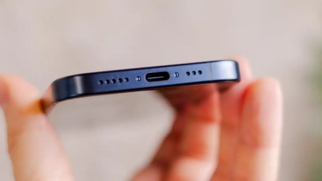 من iPhoneIslam.com، لقطة مقربة لجهاز آي فون نحيف أسود أنيق ممسوك بين الأصابع، وتظهر حافته السفلية مع منفذ USB-C مركزي وشبكات مكبر الصوت على كلا الجانبين.