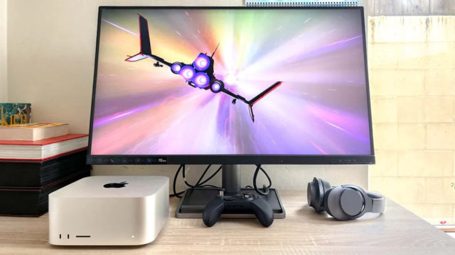 From iPhoneIslam.com, a computer screen showing a spaceship flying through space surrounded by a Mac Studio base unit, a gaming console, and headphones on a desk.