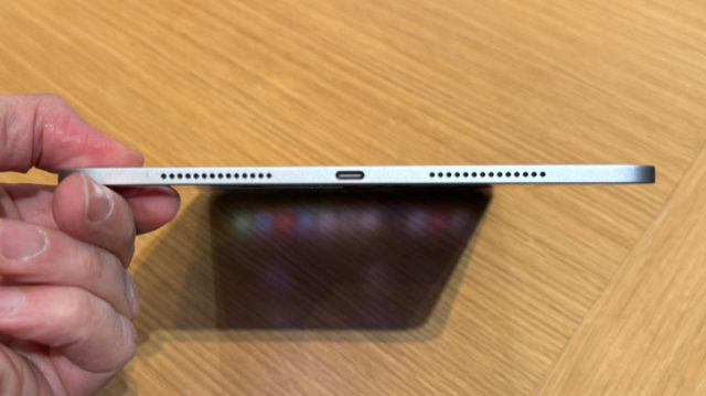 من iPhoneIslam.com، منظر جانبي مقرب لشخص يحمل جهاز آي باد برو رفيع يظهر منفذ USB-C وشبكات السماعات على خلفية سطح خشبي.