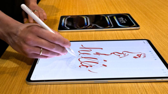 من iPhoneIslam.com، شخص يستخدم قلمًا للرسم على جهاز آي باد إير يعرض كلمة "مرحبًا" ورسمًا للوجه. يوجد جهاز لوحي آخر على الطاولة القريبة.