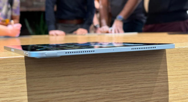 من iPhoneIslam.com، لقطة مقربة لجهاز آي باد برو ذو شكل جانبي رفيع يستقر على سطح خشبي. الخلفية غير واضحة قليلاً، وتظهر الأشخاص واقفين ومتفاعلين.