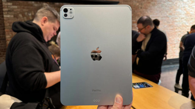 من iPhoneIslam.com، شخص يحمل جهاز Apple آي باد برو فضي اللون مع ظهور الكاميرا الخلفية، بينما ينظر الآخرون إلى الأجهزة في الخلفية.