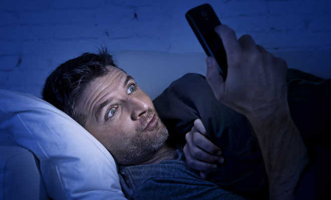 من iPhoneIslam.com، رجل يرقد في السرير تحت بطانية، ممسكًا بجهاز iPhone الخاص به وينظر إلى شاشته. الغرفة مضاءة بشكل خافت بالضوء الأزرق، مما يجعلها مثالية للنوم.