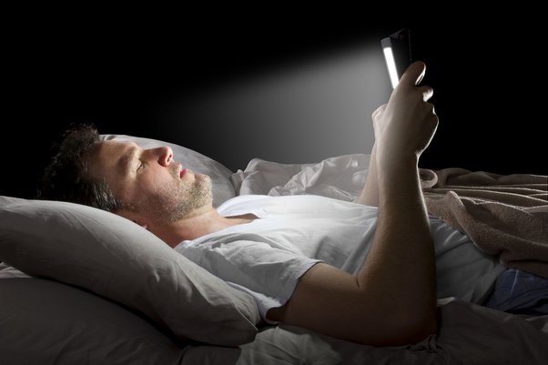 من iPhoneIslam.com، رجل يرقد على السرير في غرفة ذات إضاءة خافتة، ويمسك بجهاز لوحي ينبعث منه ضوء ساطع وينظر إليه. يفكر في التبديل إلى شاشة الآي فون من أجل تقليل الضغط عند النوم.