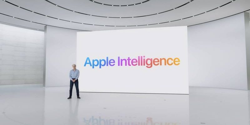 من iPhoneIslam.com، يقف فرد في غرفة بيضاء حديثة بها شاشة كبيرة تعرض كلمة "Apple Intelligence" بأحرف ملونة، ويتابع آخر الأخبار.