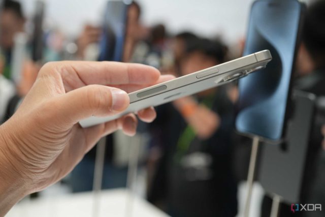 من iPhoneIslam.com، شخص يحمل الآي-فون الفضي بشكل جانبي في مكان مزدحم؛ يتم عرض الهواتف الذكية الأخرى على المدرجات في الخلفية.