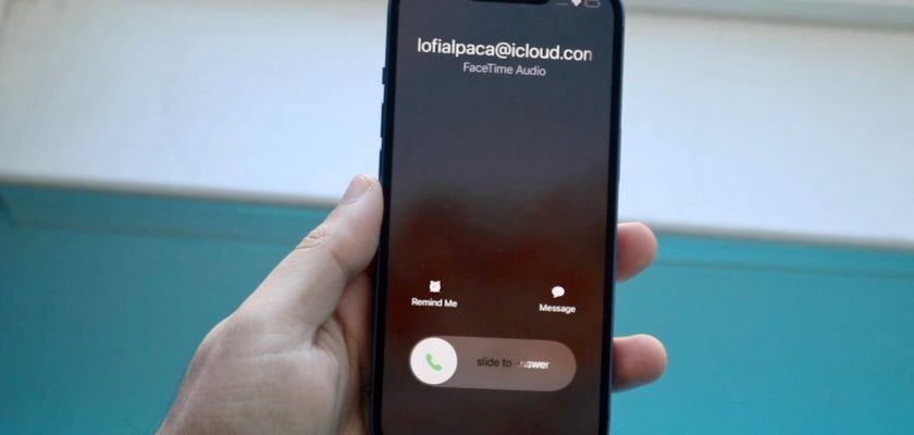 من iPhoneIslam.com، يد تحمل الآي فون تعرض مكالمة FaceTime الصوتية الواردة من "lofialpaca@icloud.com"، مع خيارات "تذكيري" أو "رسالة" أو "التمرير للرد" بينما يومض زر الباور في الخلفية، جاهزًا للاستخدام.