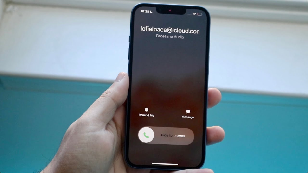 من iPhoneIslam.com، يد تحمل الآي فون تعرض مكالمة FaceTime الصوتية الواردة من "lofialpaca@icloud.com"، مع خيارات "تذكيري" أو "رسالة" أو "التمرير للرد" بينما يومض زر الباور في الخلفية، جاهزًا للاستخدام.