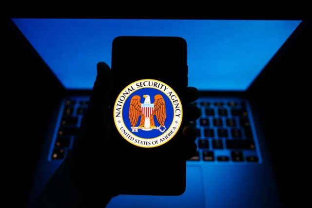من iPhoneIslam.com، يد تحمل هاتفًا ذكيًا يعرض شعار وكالة الأمن القومي (NSA)، مع تسليط الضوء على حماية الآي فون، مع توهج جهاز كمبيوتر محمول باللون الأزرق في الخلفية.