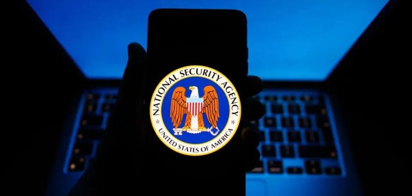 من iPhoneIslam.com، يد تحمل هاتفًا ذكيًا يعرض شعار وكالة الأمن القومي (NSA)، مع تسليط الضوء على حماية الآي فون، مع توهج جهاز كمبيوتر محمول باللون الأزرق في الخلفية.