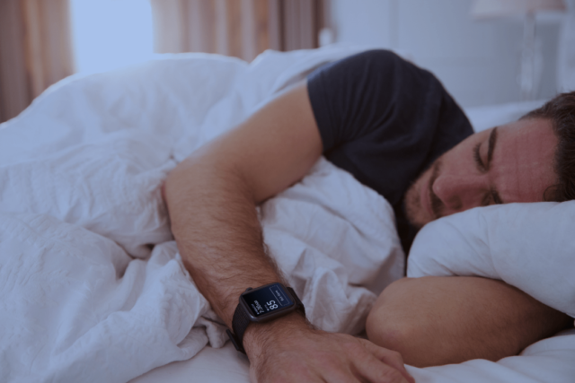 Da iPhoneIslam.com, una persona che indossa uno smartwatch Garmin è sdraiata sul letto sotto una trapunta bianca, sembra addormentata.