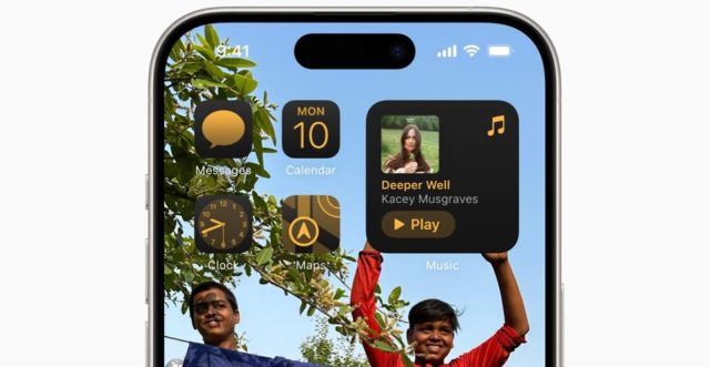 من iPhoneIslam.com، شاشة آي فون الرئيسية تعرض العديد من التطبيقات بما في ذلك الرسائل والتقويم والساعة والخرائط والموسيقى مع تشغيل أغنية "Deeper Well" من تأليف Kacey Musgraves، وصورة خلفية لطفلين مبتسمين في الهواء الطلق.