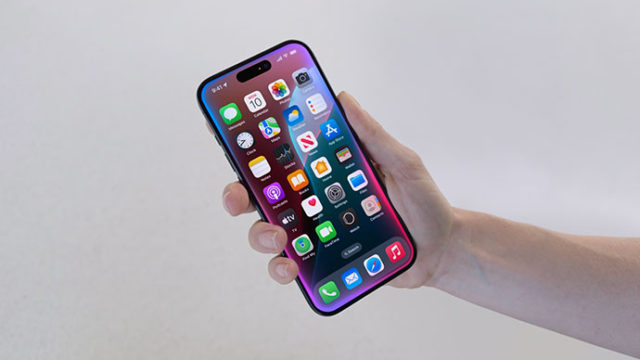من iPhoneIslam.com، يد تحمل هاتفًا ذكيًا مع أيقونات تطبيقات ملونة متنوعة معروضة على الشاشة.