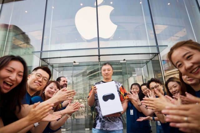 Van iPhoneIslam.com: Een man staat in juli met een doos met daarin een Apple Vision Pro buiten een Apple Store, omringd door een groep mensen die klappen en glimlachen.