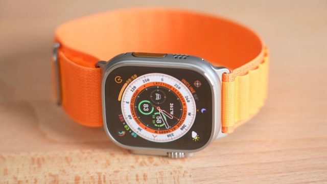 На сайте iPhoneIslam.com Apple Watch изображены с оранжевой полосой на деревянной поверхности. На циферблате отображаются различные показатели, включая время, уровень заряда батареи и информацию о компасе. Эта конкретная модель — Apple Altra 3.