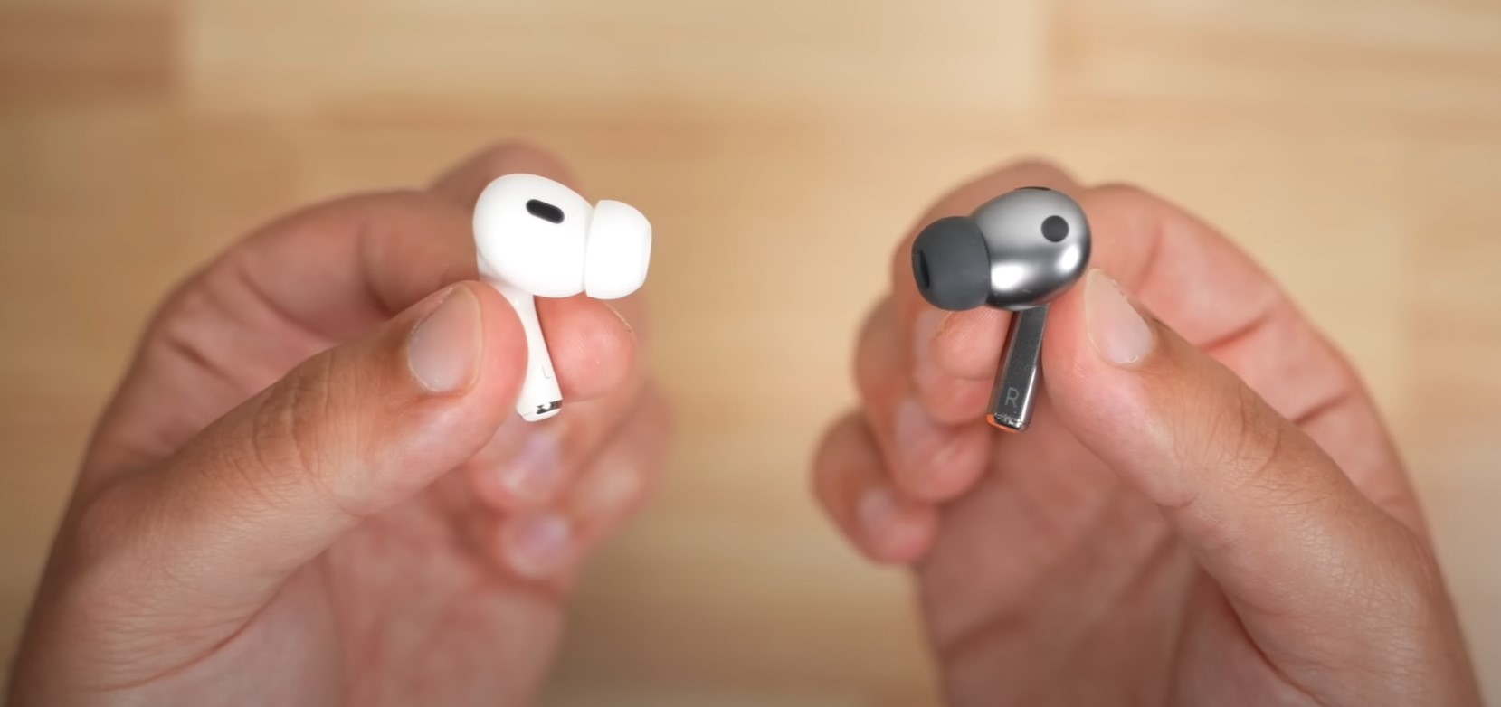 من iPhoneIslam.com، يدان تحملان سماعات أذن لاسلكية؛ تحمل يد واحدة سماعة أذن بيضاء اللون AirPods Pro 2، بينما تحمل اليد الأخرى سماعة أذن رمادية اللون من شركة Samsung Buds3 pro. الخلفية غير واضحة.