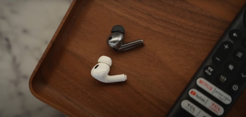 من iPhoneIslam.com، سماعتان لاسلكيتان، Samsung Buds3 pro باللون الأسود وAirPods pro 2 باللون الأبيض، موضوعتان على سطح خشبي بجوار جهاز التحكم عن بعد باللون الأسود.