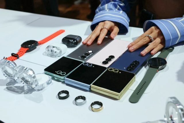 Da iPhoneIslam.com, tre smartphone con vari smartwatch Samsung, auricolari e accessori disposti su una superficie bianca.