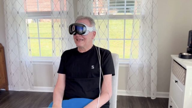 From iPhoneIslam.com A person sits indoors, wearing a wired virtual reality headset, in a room with large windows and sheer white curtains, likely watching sideline news from the week of July 26.