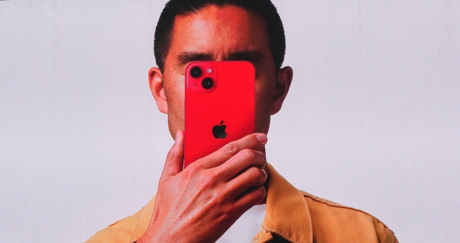 من iPhoneIslam.com، شخص يحمل هاتفًا ذكيًا أحمر اللون عليه شعار Apple أمام وجهه، ويرتدي سترة بيج على خلفية عادية. احذر منها علامات تشير إلى احتمال حدوث خطأ في جهاز الكمبيوتر.
