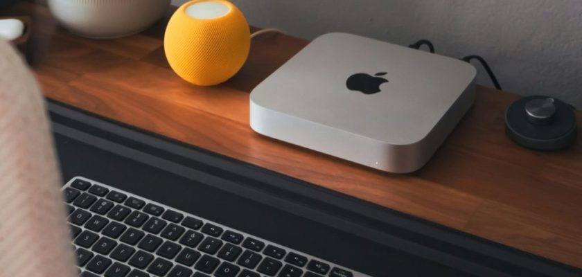 من iPhoneIslam.com، يوضع جهاز Mac mini ولوحة مفاتيح لاسلكية ومكبر صوت أصفر صغير بشكل أنيق على المكتب الخشبي.