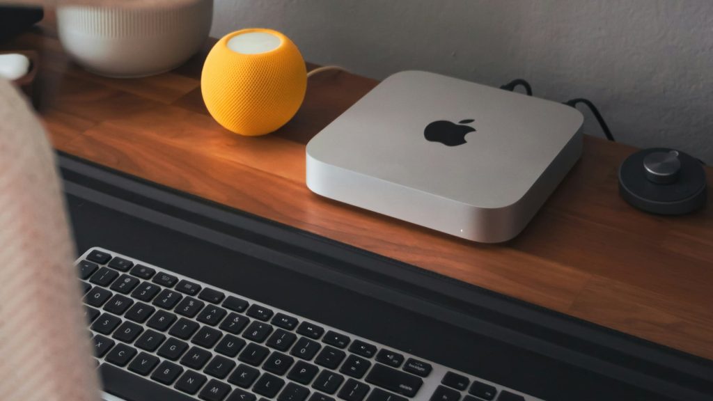 من iPhoneIslam.com، يوضع جهاز Mac mini ولوحة مفاتيح لاسلكية ومكبر صوت أصفر صغير بشكل أنيق على المكتب الخشبي.