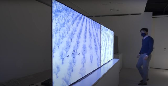 From iPhoneIslam.com A masked man stands next to two large flat screen TVs showing aerial images of a snow-covered landscape with rows of trees, anticipating the launch of the iPhone 16 in one month.