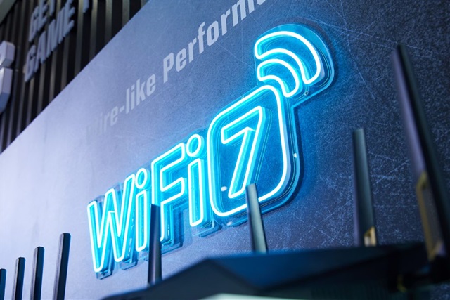 From iPhoneIslam.com An illuminated sign displaying “WiFi 7” in blue neon lights catches your eye, with the top of several router antennas subtly appearing in the foreground.