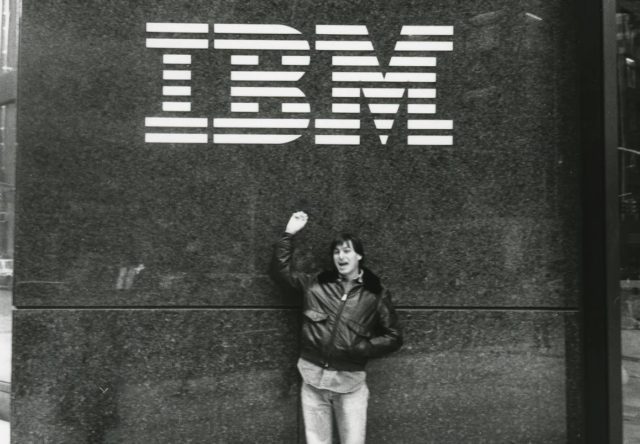 Depuis iPhoneIslam.com Une personne vêtue d'une veste se tient à côté du mur portant le logo IBM, lève une main en souriant, capturant un moment pendant l'actualité en marge.