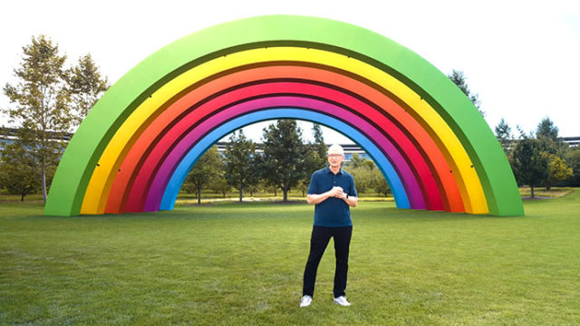 Depuis iPhoneIslam.com Une personne se tient sur l’herbe devant une grande structure arc-en-ciel colorée, avec des arbres en arrière-plan.