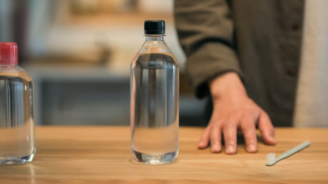 Desde iPhoneIslam.com, una persona se para frente a una mesa de madera con una botella de agua transparente, una botella con tapa roja y un palo cerca, y limpia cuidadosa y meticulosamente los AirPods.