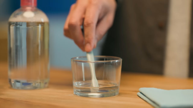 From iPhoneIslam.com, A person stirs liquid in a cup using a stick, next to a bottle of apple juice and a cleaning cloth on a wooden surface.