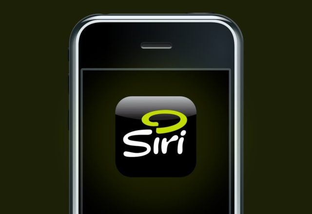 من iPhoneIslam.com، صورة مقربة لشاشة هاتف ذكي تعرض أيقونة تطبيق Siri، والتي تحتوي على حرف 'S' منمق مع لمسة من اللون الأخضر النيون على خلفية سوداء، مما يشير إلى نشأة Siri وتطورها في التكنولوجيا الحديثة.