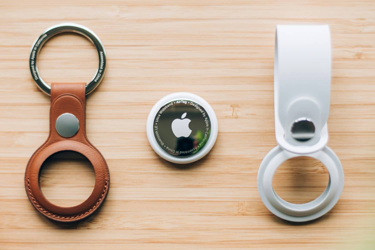 iPhoneIslam.com에서 왼쪽에 갈색 가죽 키홀더가 있고 오른쪽에 흰색 키홀더가 있는 Apple AirTag 2가 나무 표면 위에 우아하게 놓여 있습니다.