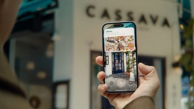 من iPhoneIslam.com، شخص يحمل هاتفًا ذكيًا يعمل بنظام التشغيل iOS 18.2، ويعرض ملفًا شخصيًا عبر الإنترنت لمطعم وصورًا على خلفية مبنى مكتوب عليه "Cassava"، مما يسلط الضوء على قدرات الذكاء البصري المحسنة للمنصة.
